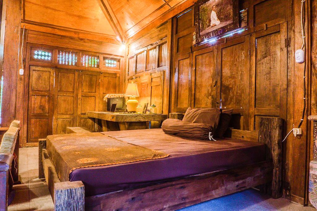 A cozy sleeping area features wooden walls and ceiling, creating a warm ambiance. A spacious bed with dark bedding is positioned centrally, complemented by rustic wooden furniture. Soft lighting emanates from a lamp on a nearby table, adding to the calming atmosphere.