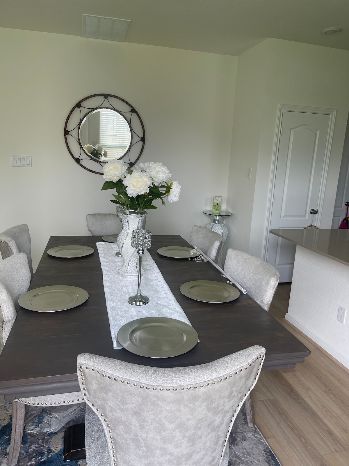 A dining area features a long wooden table set for six, accented by simple dinnerware in soft tones. A decorative vase filled with white flowers stands at the center. A round mirror adorns the wall, enhancing the ambient light in the space.