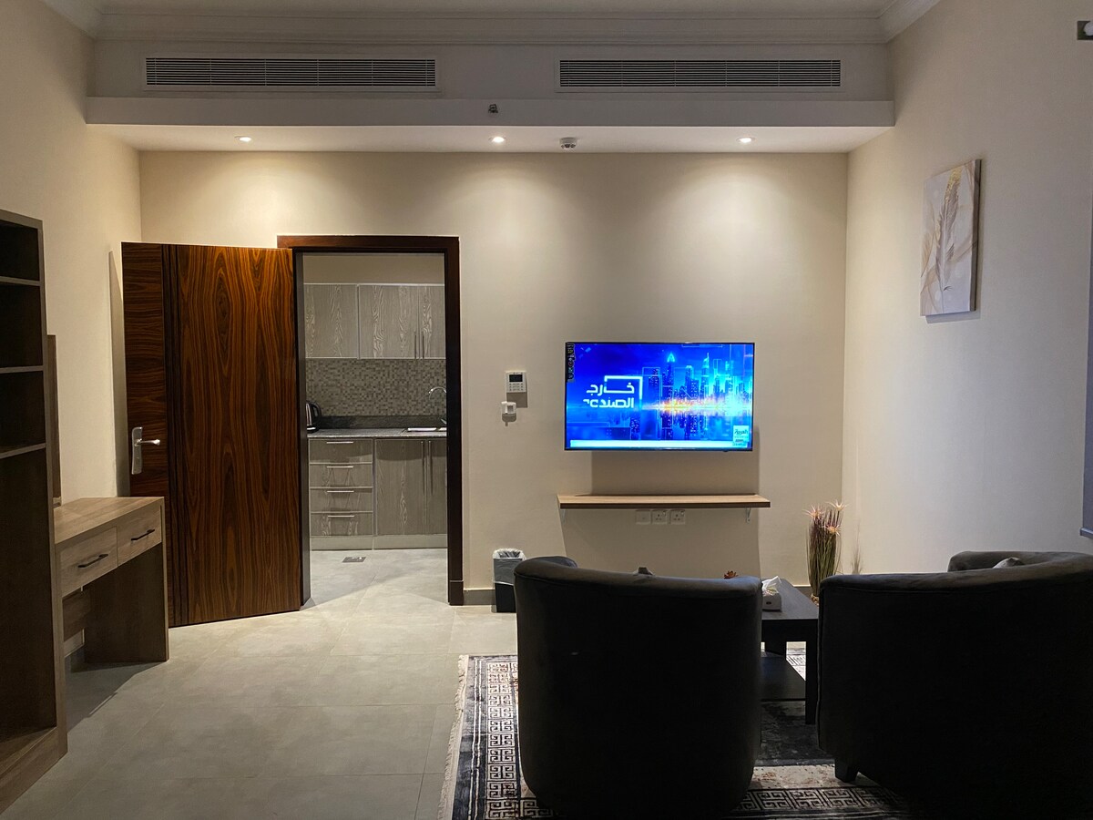 A modern living area is highlighted by a wall-mounted television, facing two comfortable chairs. A wooden desk and a small kitchen area are visible in the background. Soft lighting contributes to a calm atmosphere, with gray tiles underfoot and a decorative rug adding texture.