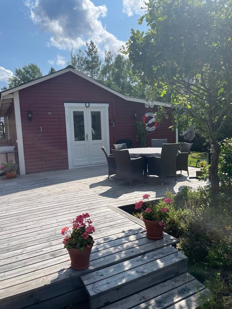 A red building with a white door is featured surrounded by greenery. A wooden deck leads to a circular table set with chairs. Potted flowers are placed at the entrance, and the warm sunlight casts shadows on the decking.