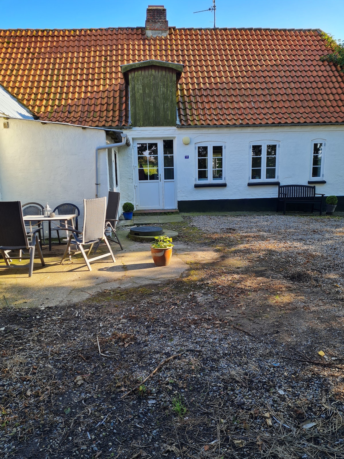 A quaint outdoor area features a gravel patio with a round dining table surrounded by six chairs. A small flower pot rests on the ground, while a wooden bench contrasts with the white exterior of the house. Sunlight casts gentle shadows on the scene.