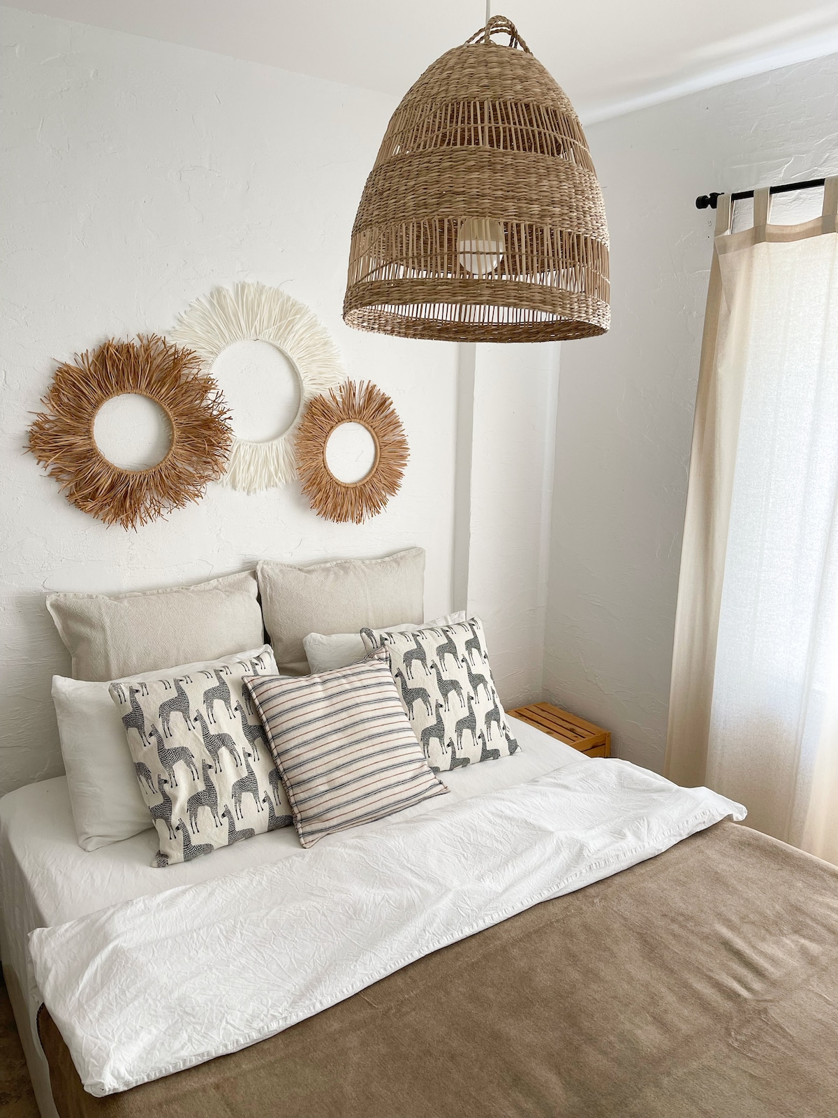 A serene bedroom arrangement features a spacious bed adorned with a light-colored blanket. Decorative cushions with varied patterns are arranged on the bed. Wall art consisting of circular mirrors and natural materials is displayed above the headboard, with soft curtains framing a nearby window.