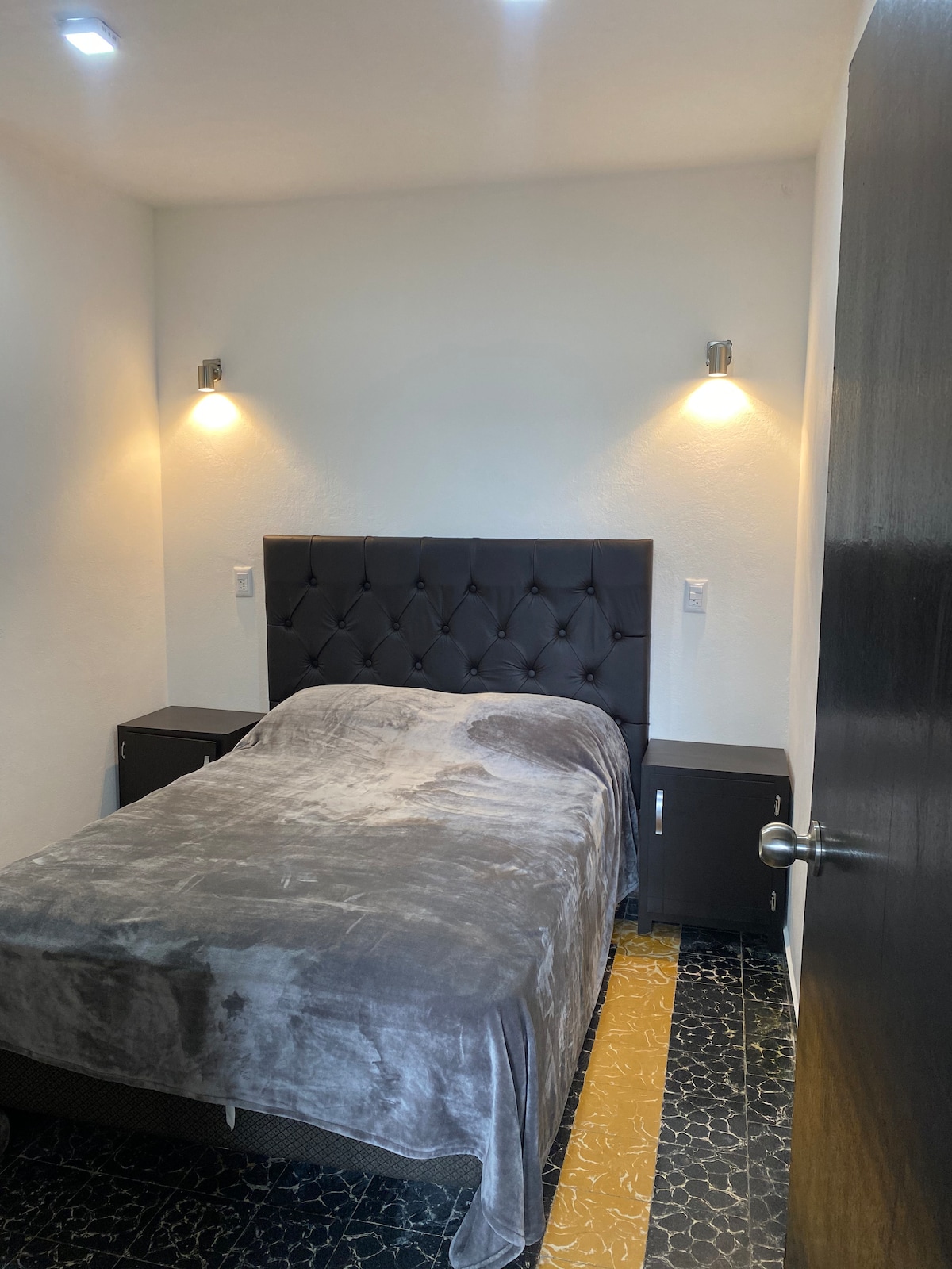 A cozy sleeping area is presented with a spacious bed covered by a soft, neutral blanket. Two bedside tables with modern lamps are positioned on either side of the headboard, providing gentle lighting. The room features a simple yet functional design, enhanced by light-colored walls.