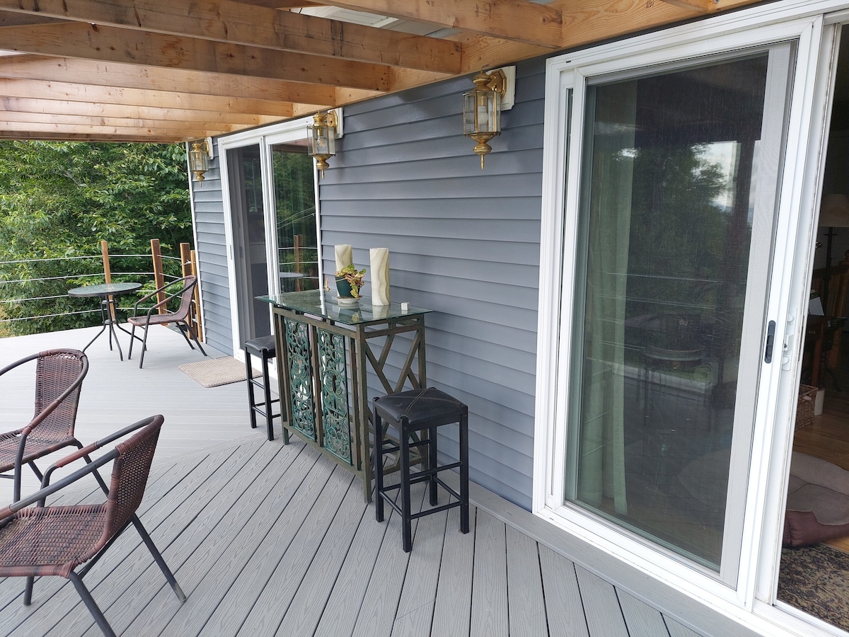A spacious deck features outdoor seating options, including two chairs and a bar-height table with stools. The deck is partially covered, providing shade, while large sliding glass doors lead into the living area, allowing natural light to flow inside.
