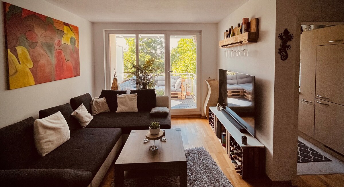A spacious living area features a large sectional sofa adorned with various cushions. Natural light pours in through expansive glass doors, providing a view of greenery outside. A modern television is positioned on a console table, and a vibrant piece of art adds color to the wall.