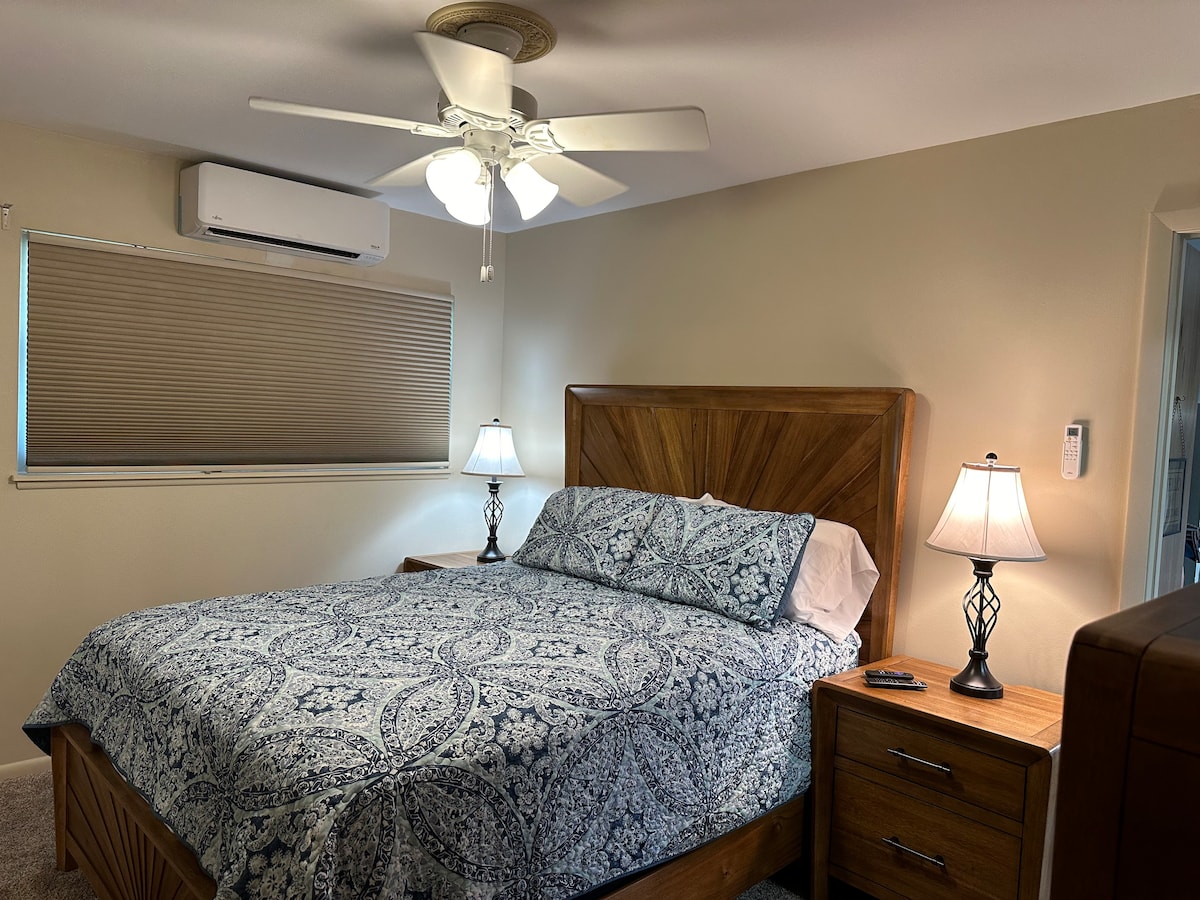 A comfortable bedroom is featured with a spacious bed covered in a patterned blue quilt. Two bedside lamps provide soft lighting on either side of the bed, and an air conditioning unit is mounted above a window with a light-filtering shade.