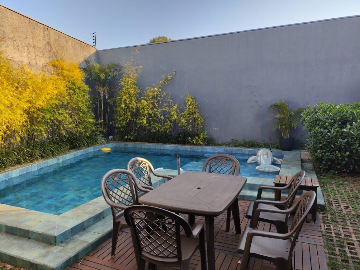 A private pool area features clear water surrounded by lush greenery and a modern gray wall. A wooden table with several chairs is placed nearby, providing a space for outdoor dining and relaxation.