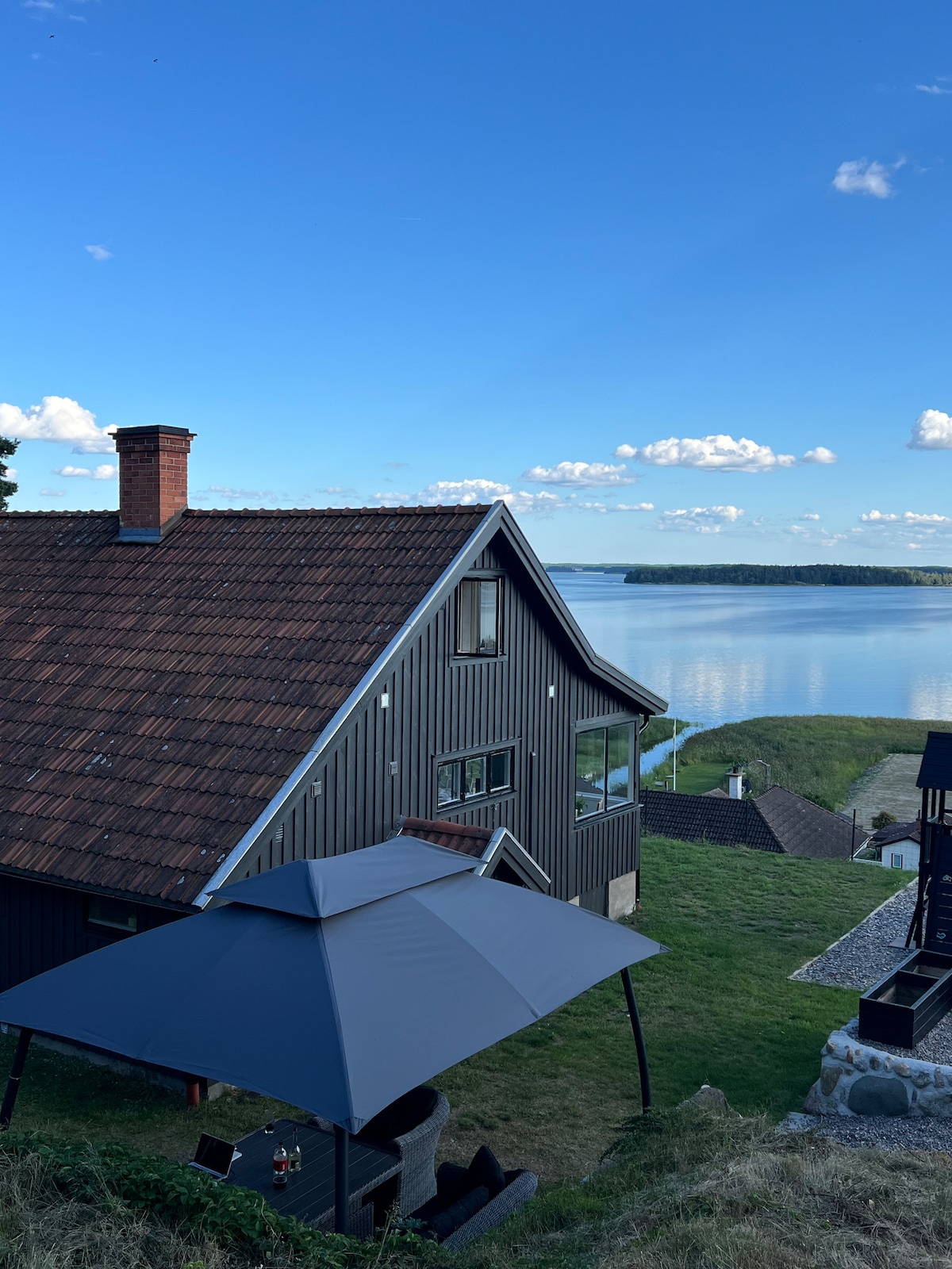 A charming wooden house with a shingled roof is positioned on a hillside, overlooking a calm body of water. A grey umbrella provides shade over a seating area, while large windows reflect the serene landscape and sky, creating a connection between indoors and outdoors.