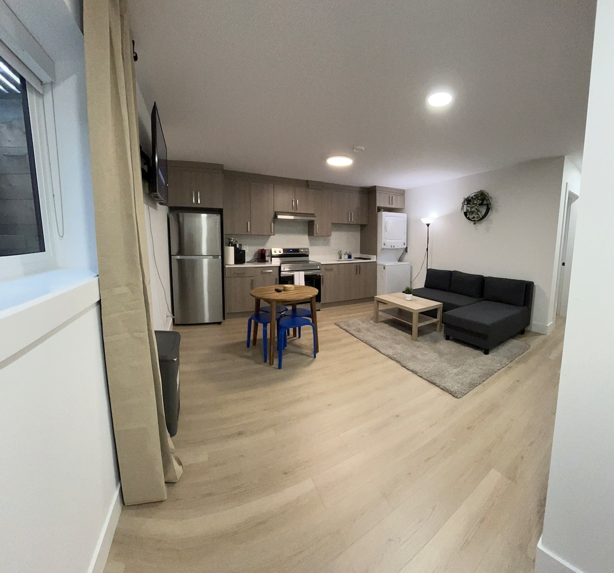 An open-plan living area is shown, featuring a modern grey sofa and a small wooden table with blue chairs. A fully-equipped kitchen is visible with stainless steel appliances, including a stove and refrigerator. Natural light enters through a nearby window, complemented by light wood flooring.