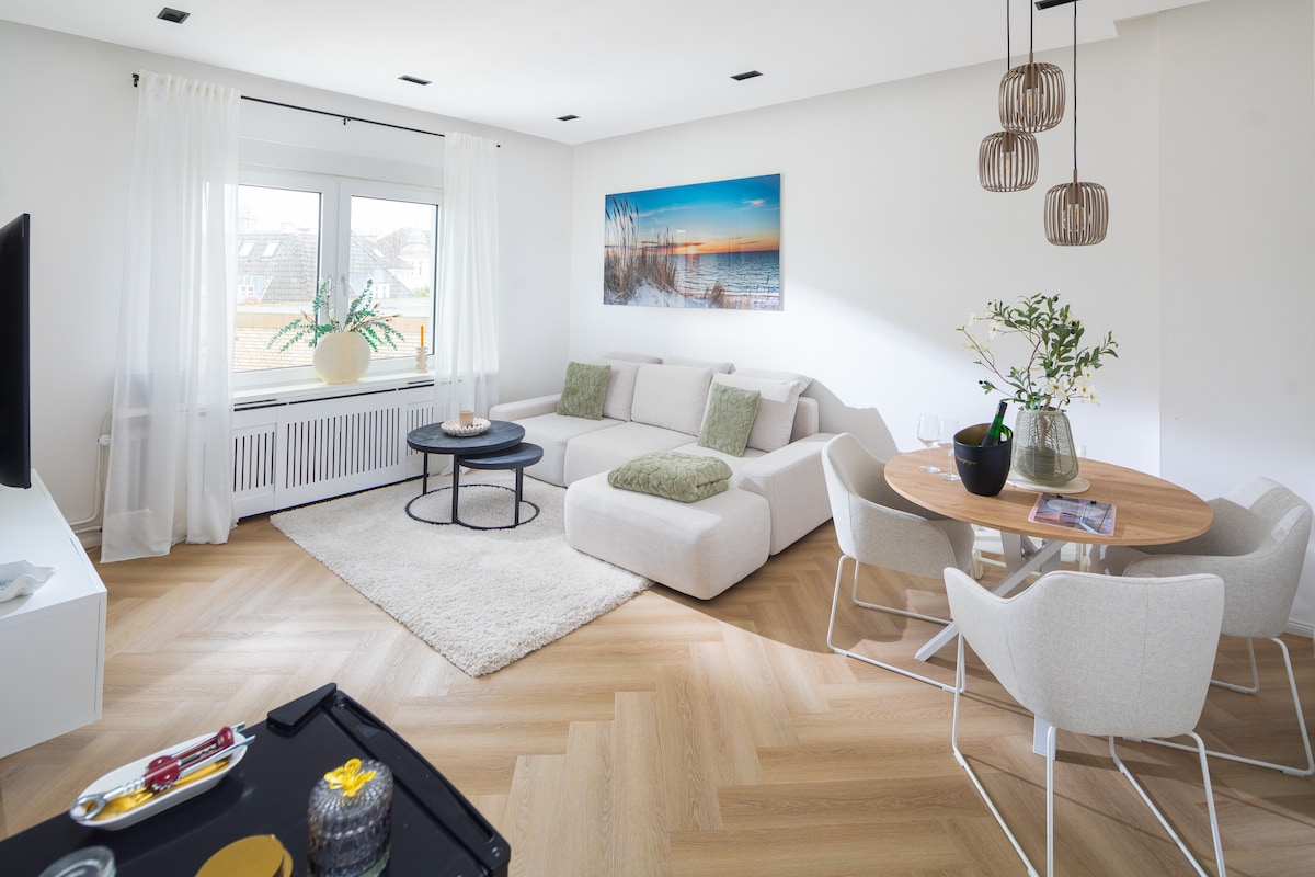 A bright living area features a white sectional sofa adorned with decorative pillows. A round coffee table sits in front of the sofa, with a soft area rug underneath. A dining table with light-colored chairs is positioned nearby. A large window allows natural light to fill the room.
