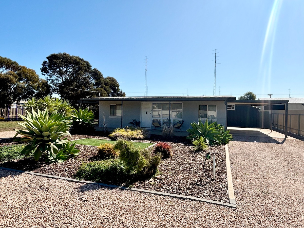 A single-story building is set against a clear blue sky, surrounded by a gravel yard featuring low-maintenance landscaping. Various plants, including succulent species, create a colorful border around the property, which is designed for easy access and relaxation.