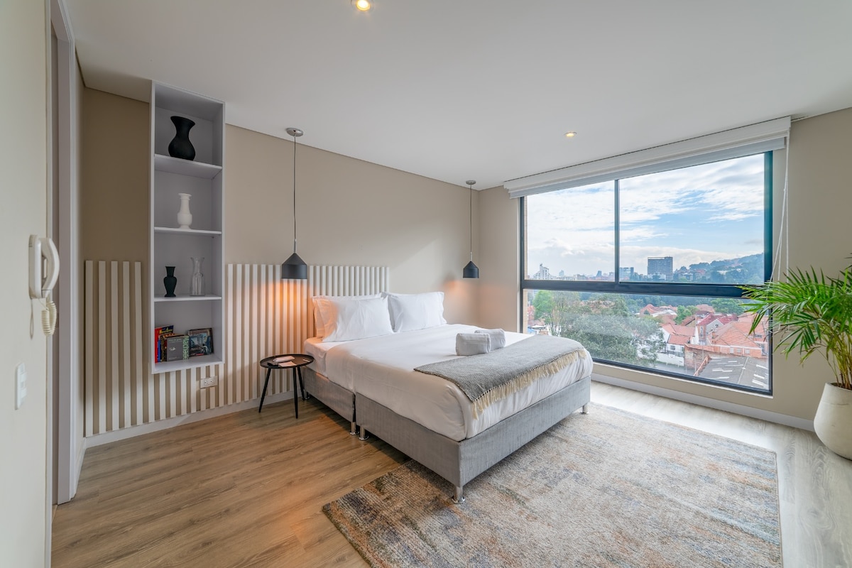 A spacious bedroom features a comfortable Queen bed adorned with neutral linens. Large windows offer a view of the cityscape, allowing natural light to fill the room. Two pendant lamps hang above nightstands, and a decorative bookshelf adds a touch of style to the area.