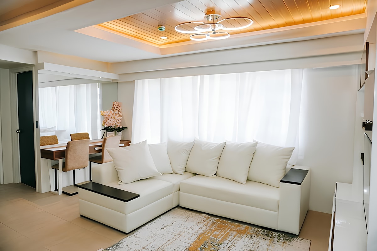 A modern living area is presented with a plush, L-shaped white sectional couch adorned with soft, white pillows. Natural light filters through sheer window treatments, illuminating the space, which features a dining area in the background with a wooden table and upholstered chairs.