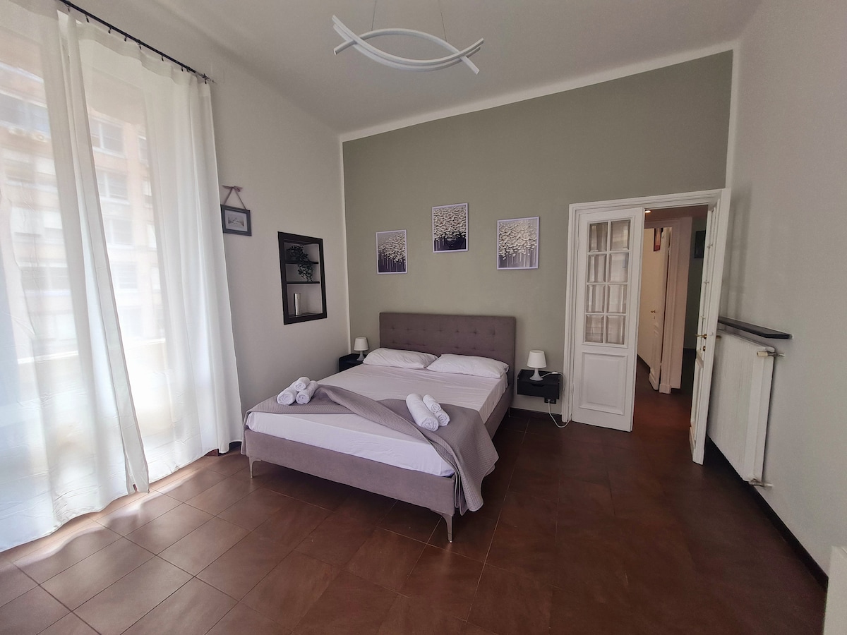 A spacious bedroom features a comfortable bed with a light duvet and decorative towels arranged neatly. Large windows allow natural light to fill the room, complemented by soft drapes. Wall art adds a touch of elegance, while the room maintains a neutral color palette.