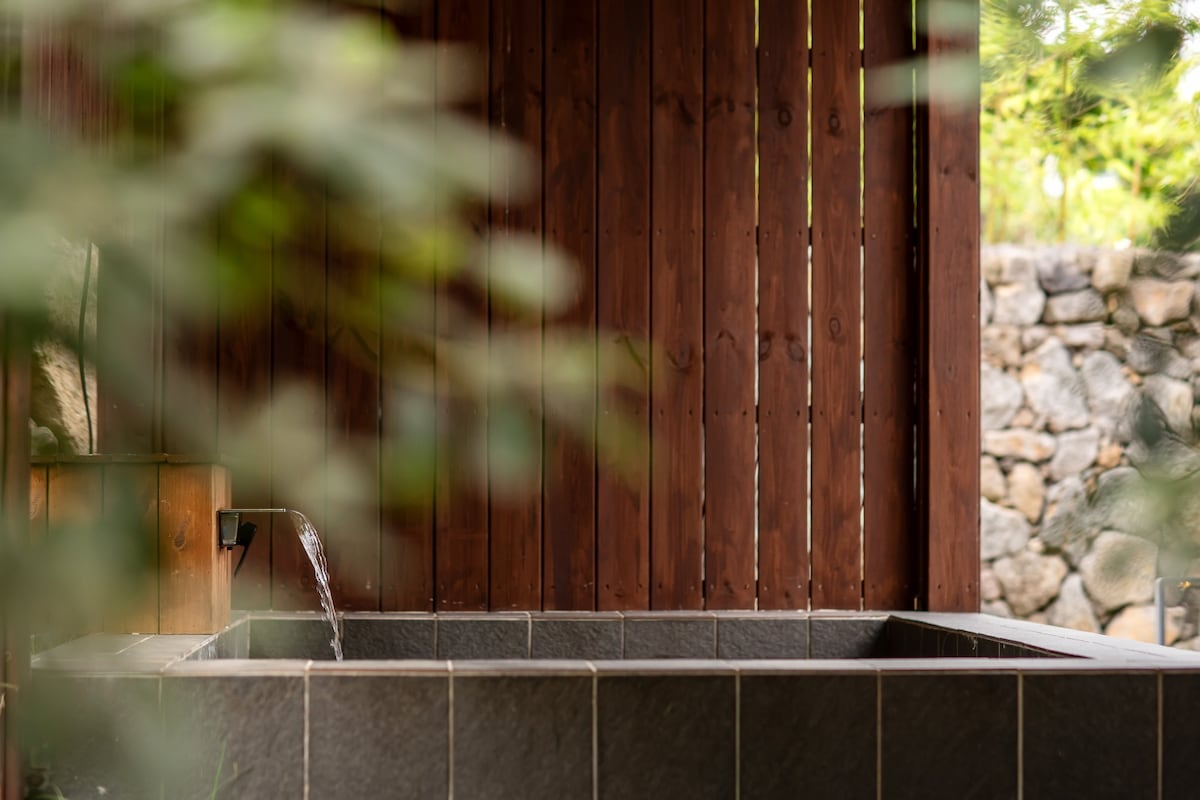 A serene outdoor tub is surrounded by wooden slats and nature. Water gently flows from a spout into the stylishly designed bath, contrasting with the natural stone wall in the background. Green foliage outlines the space, adding to the tranquil ambiance.