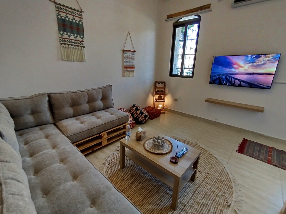 A cozy living room features a sectional sofa and a coffee table placed on a woven rug. Natural light streams in through a window, illuminating decorative wall hangings. A smart TV is mounted on the wall, providing entertainment options.