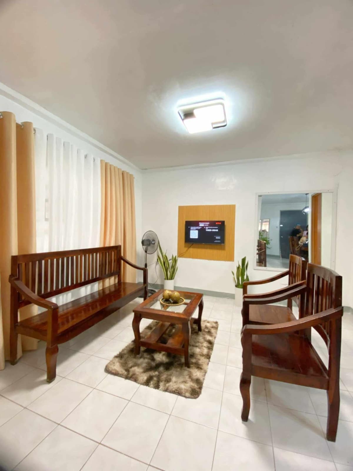 A welcoming living area features two wooden benches facing a small coffee table, which is adorned with decorative items. A fan stands nearby, and a mounted television is visible on the wall. Soft beige curtains complement the room's neutral tones.