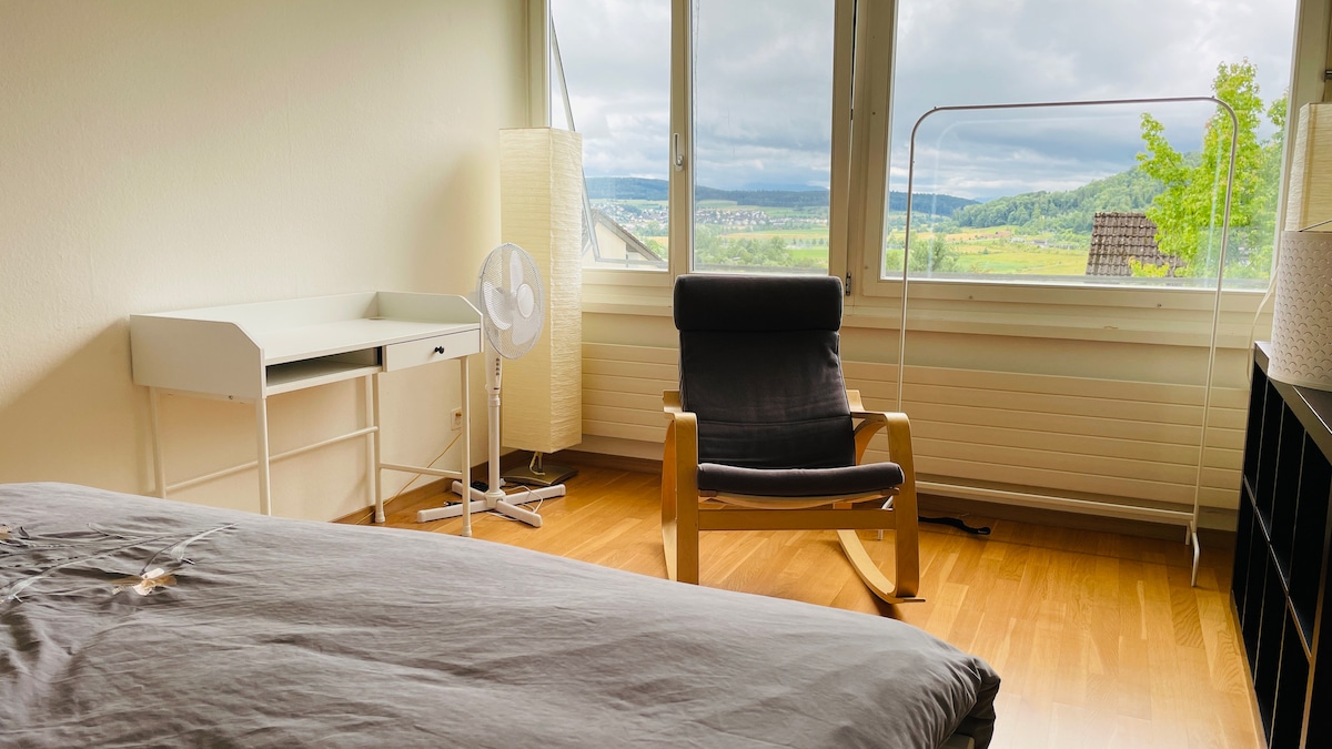 The bright bedroom features large windows offering views of the surrounding valley. A spacious bed is complemented by a wooden rocking chair and a simple desk. Light-colored walls enhance the room's airy feel, while a fan provides comfort during warmer days.
