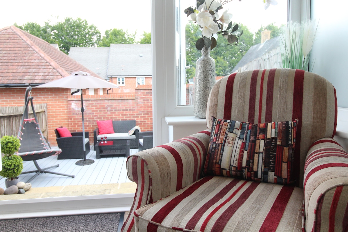 A comfortable armchair with striped upholstery sits by a large window, offering a view of a patio area furnished with a shaded table and chairs. Decorative elements include a vase with artificial flowers and a small potted plant, enhancing the inviting atmosphere.