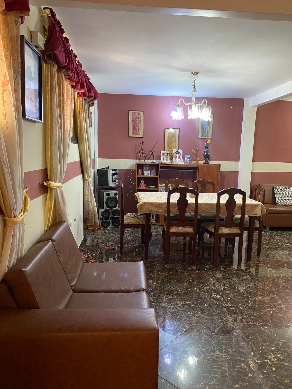 The interior area features a dining table surrounded by wooden chairs, with a decorative chandelier overhead. Brown leather seating is positioned near the front, with wall decorations adding warmth to the space. A media unit and additional furniture are visible in the background.