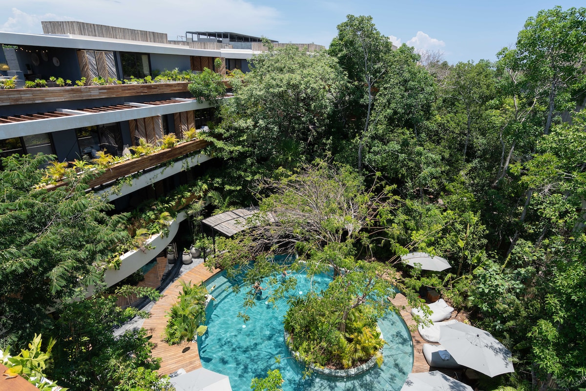 A lush green landscape surrounds a tranquil swimming pool, partially shaded by trees. Several modern lounge chairs are positioned near the pool, offering relaxation spaces. The multi-level building features balconies adorned with tropical plants, blending harmoniously with the natural environment.