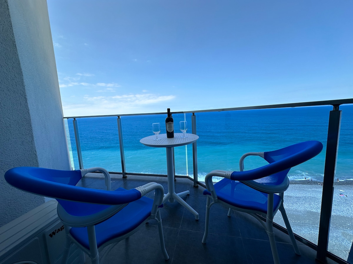 A balcony overlooks the sea, featuring a small round table with two blue chairs. A bottle of wine and two glasses are placed on the table, inviting outdoor relaxation with a scenic coastal view in the background.