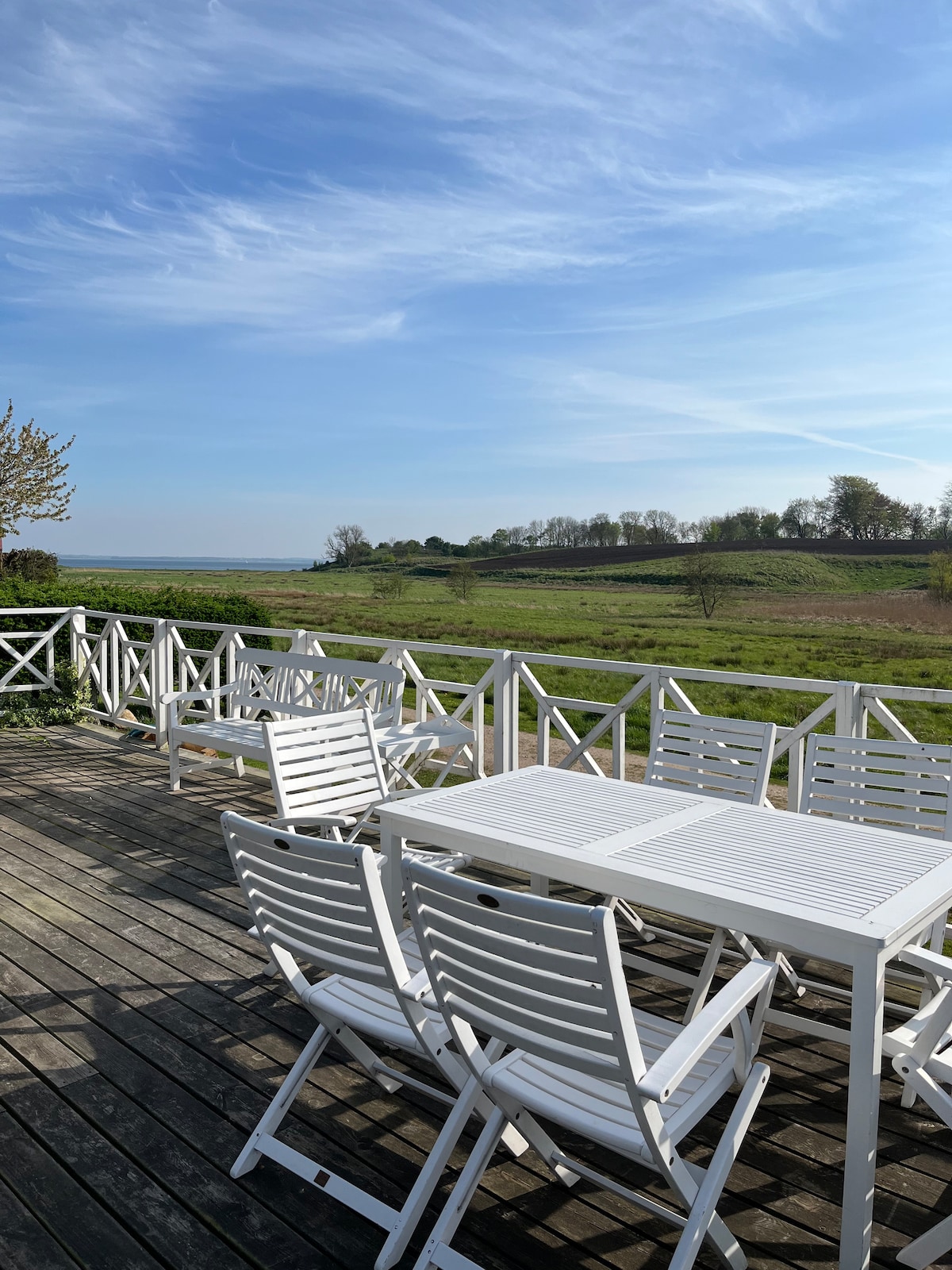 A spacious wooden terrace is furnished with white folding chairs and a matching table. The view reveals rolling green hills and scattered trees, all under a clear blue sky. The area is designed for relaxation in a natural setting.