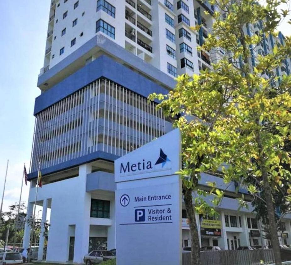 The exterior of Metia Apartment is shown, featuring a modern multi-story building with a prominent sign indicating the main entrance and parking areas for visitors and residents. A clear blue sky and greenery are visible in the foreground.