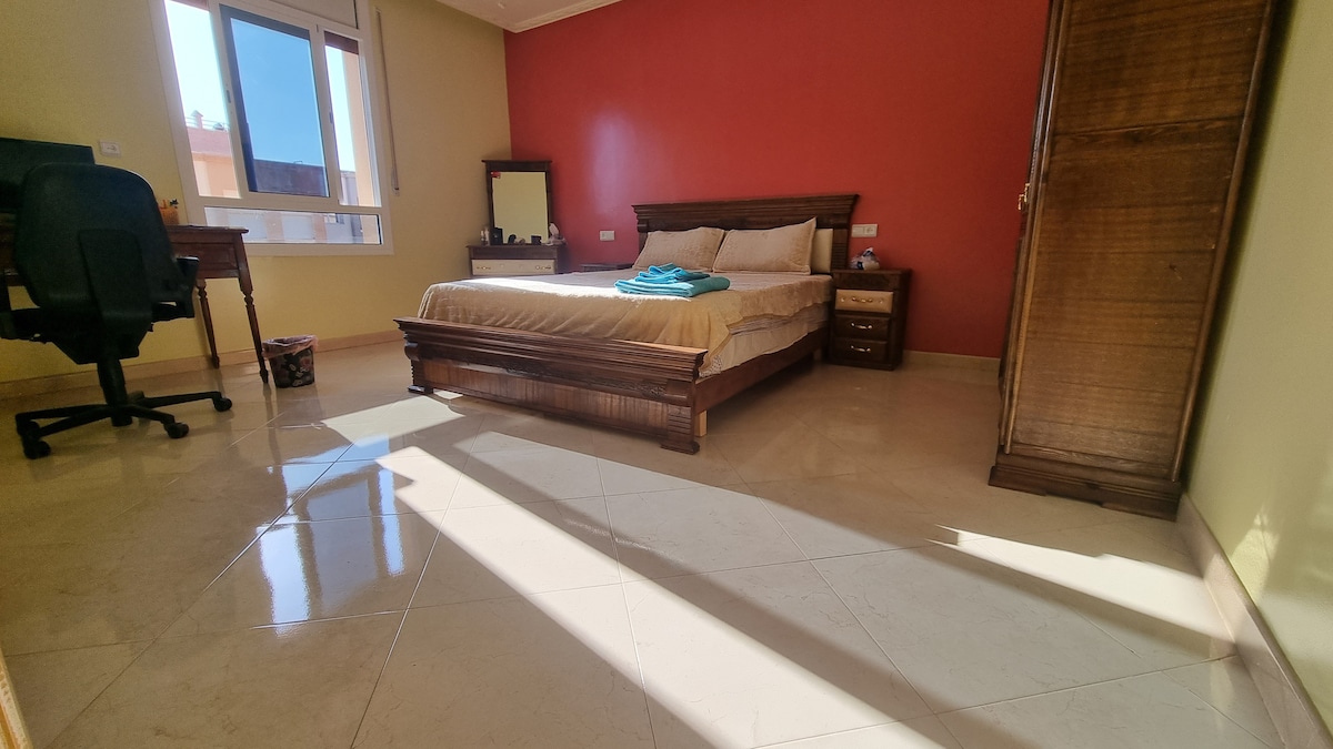A spacious bedroom is presented with a wooden bed and matching furniture, including a nightstand and a wardrobe. Natural light floods the room from large windows, creating soft shadows on the polished floor. A work desk is positioned nearby, offering a dedicated workspace.