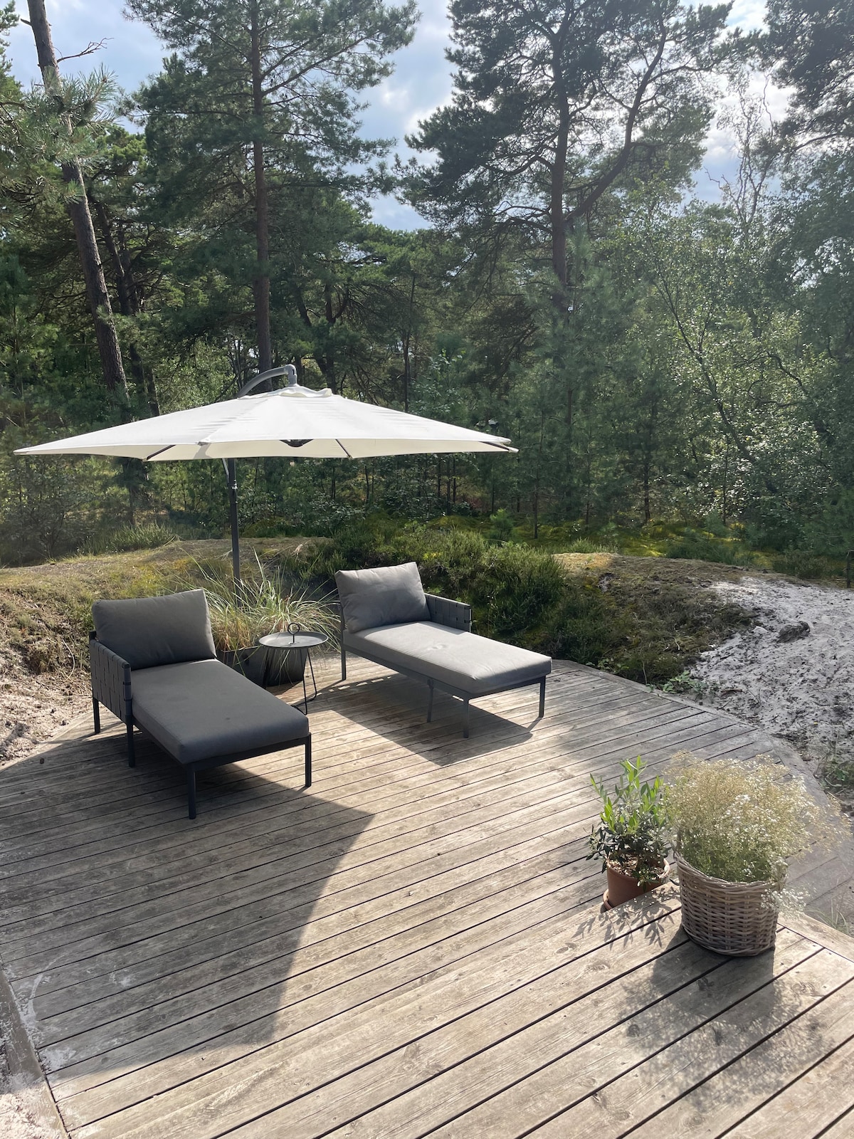 A wooden terrace is furnished with two lounge chairs and a small table, shaded by a large umbrella. Surrounding greenery creates a peaceful natural setting, providing a serene spot for relaxation. Potted plants add a touch of nature to the outdoor space.