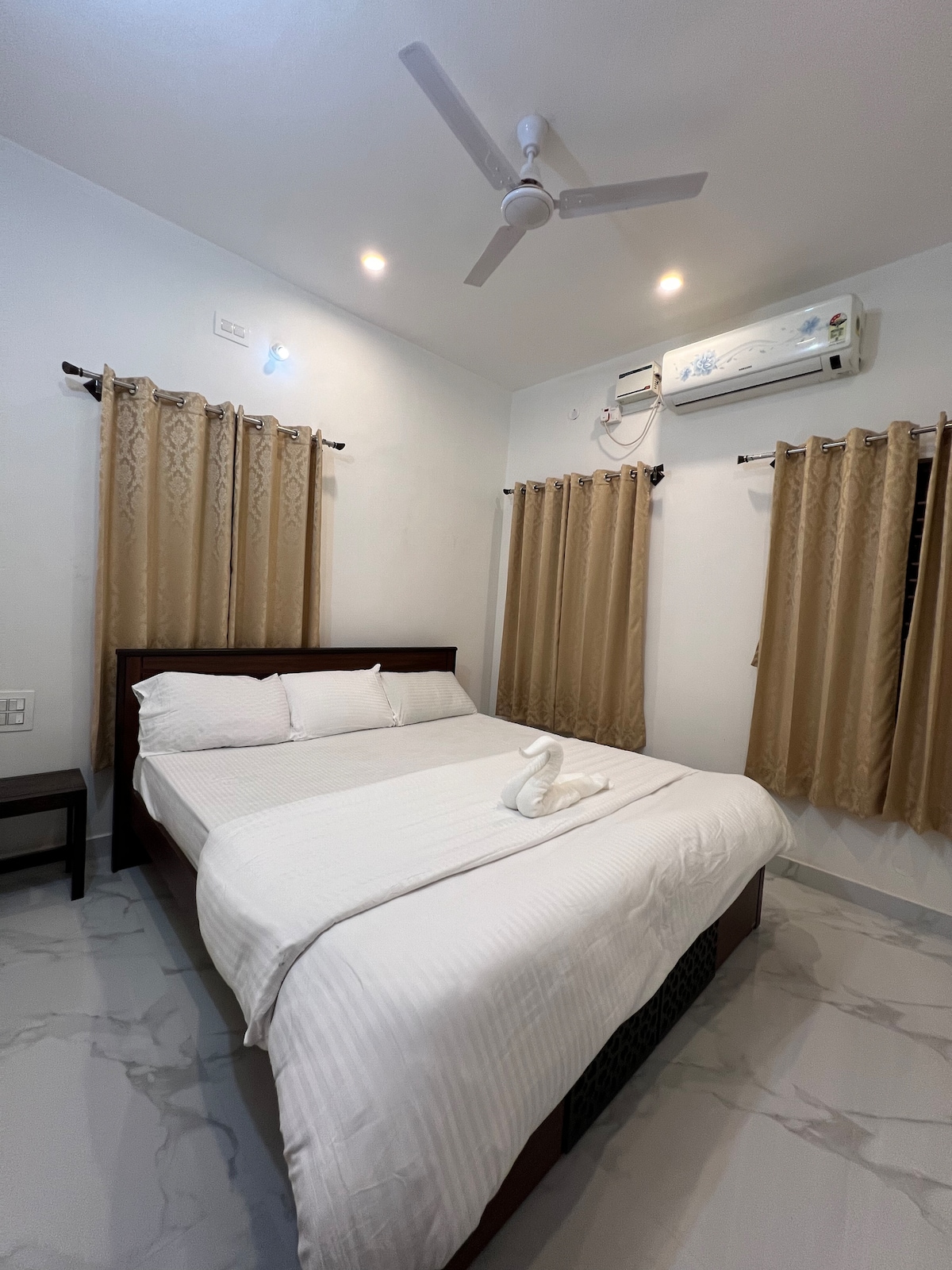 A cozy bedroom features a spacious bed dressed in white linens, with decorative towels arranged in a swan shape. Two sets of light curtains frame the windows, allowing natural light to filter in. A ceiling fan and air conditioning unit provide comfort.