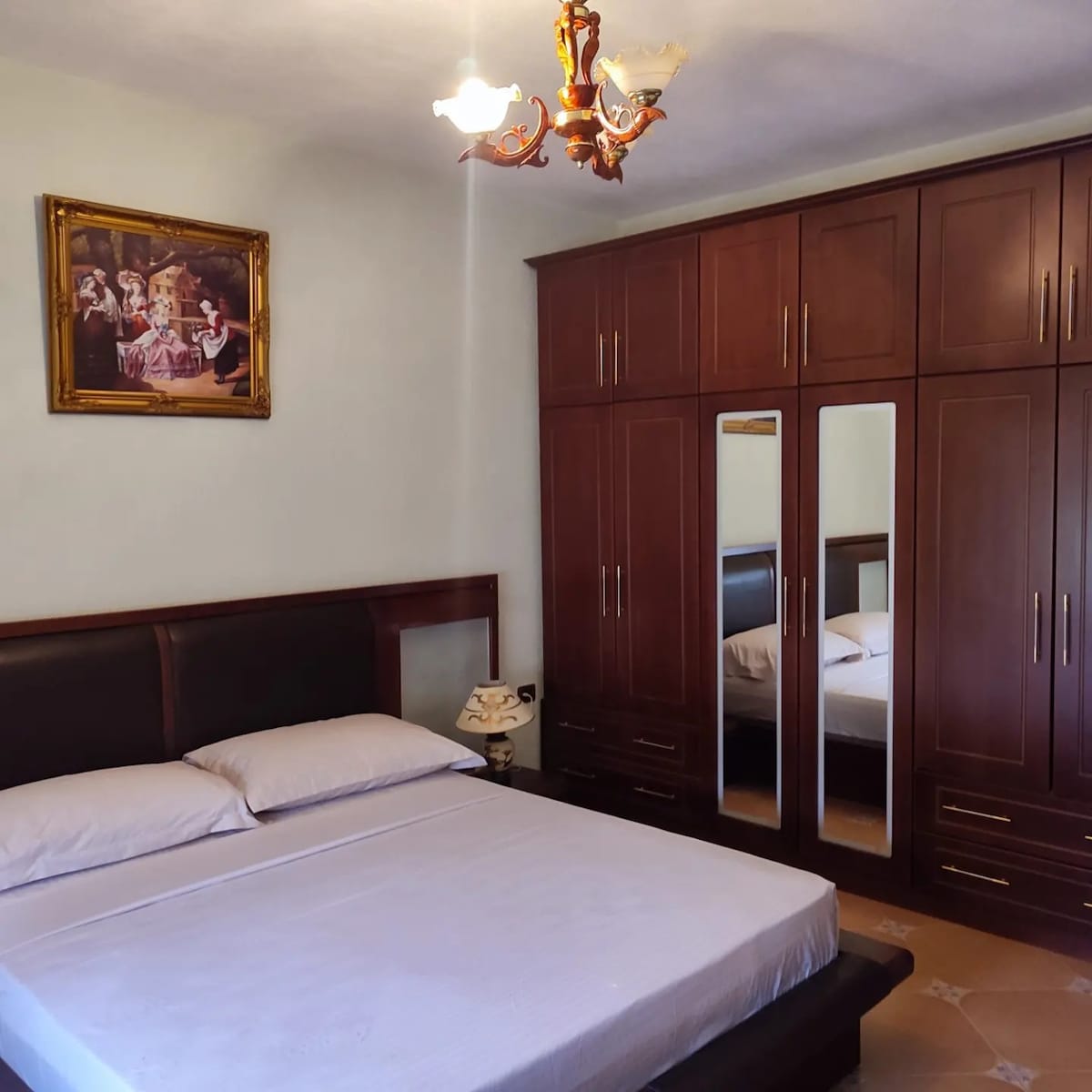 A bedroom features a spacious bed with a light-colored sheet, complemented by a wooden headboard. A large wardrobe with mirrored doors sits against the wall, reflecting the room's decor, which includes a framed painting above the bed and a bedside lamp.