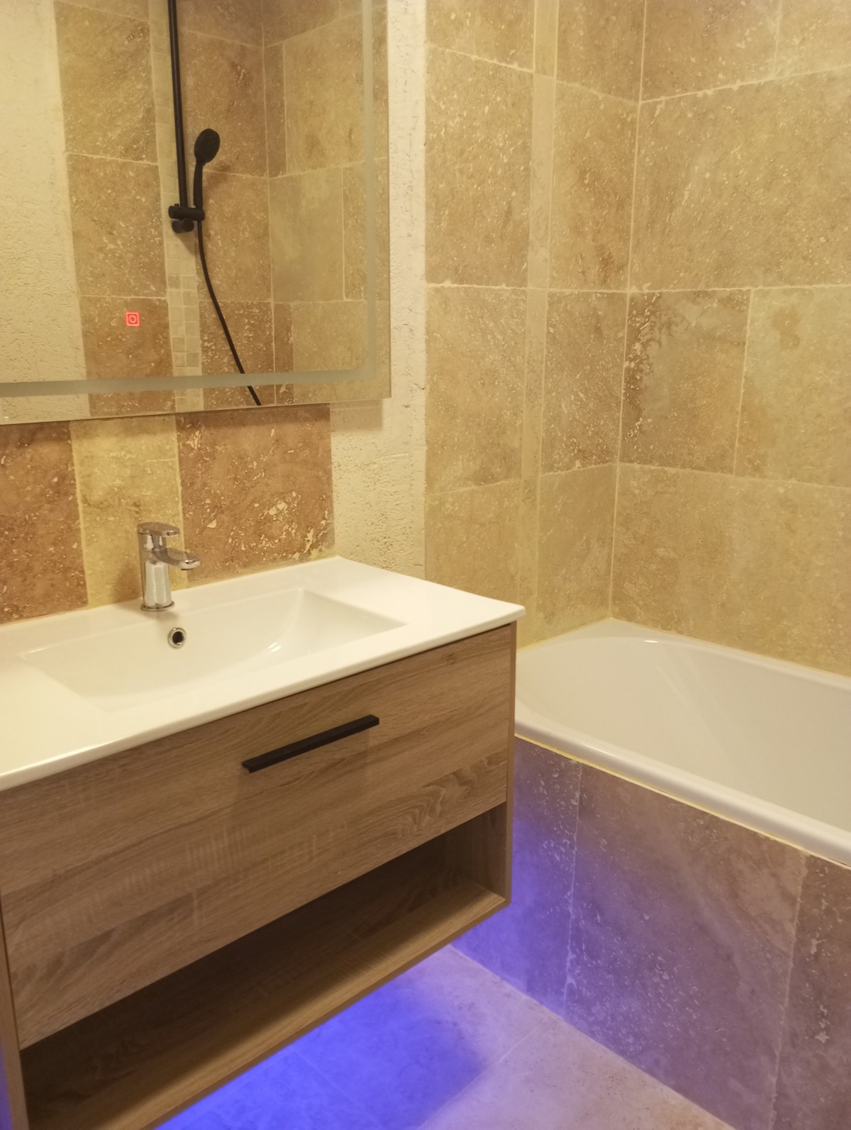 A well-appointed bathroom features a modern sink with a wooden vanity, complemented by a bathtub adjacent to the sink. Neutral-toned stone tiles cover the walls, contributing to a clean and bright atmosphere. A large mirror reflects light, enhancing the space's openness.