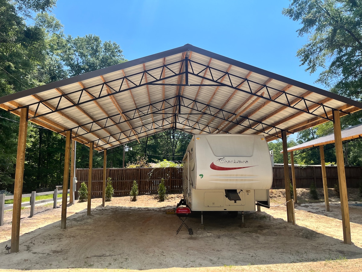 A covered parking area is seen, featuring a spacious structure supported by wooden beams. An RV is positioned beneath the roof, with a small grill set nearby on the sandy ground. Surrounding greenery provides a natural backdrop.