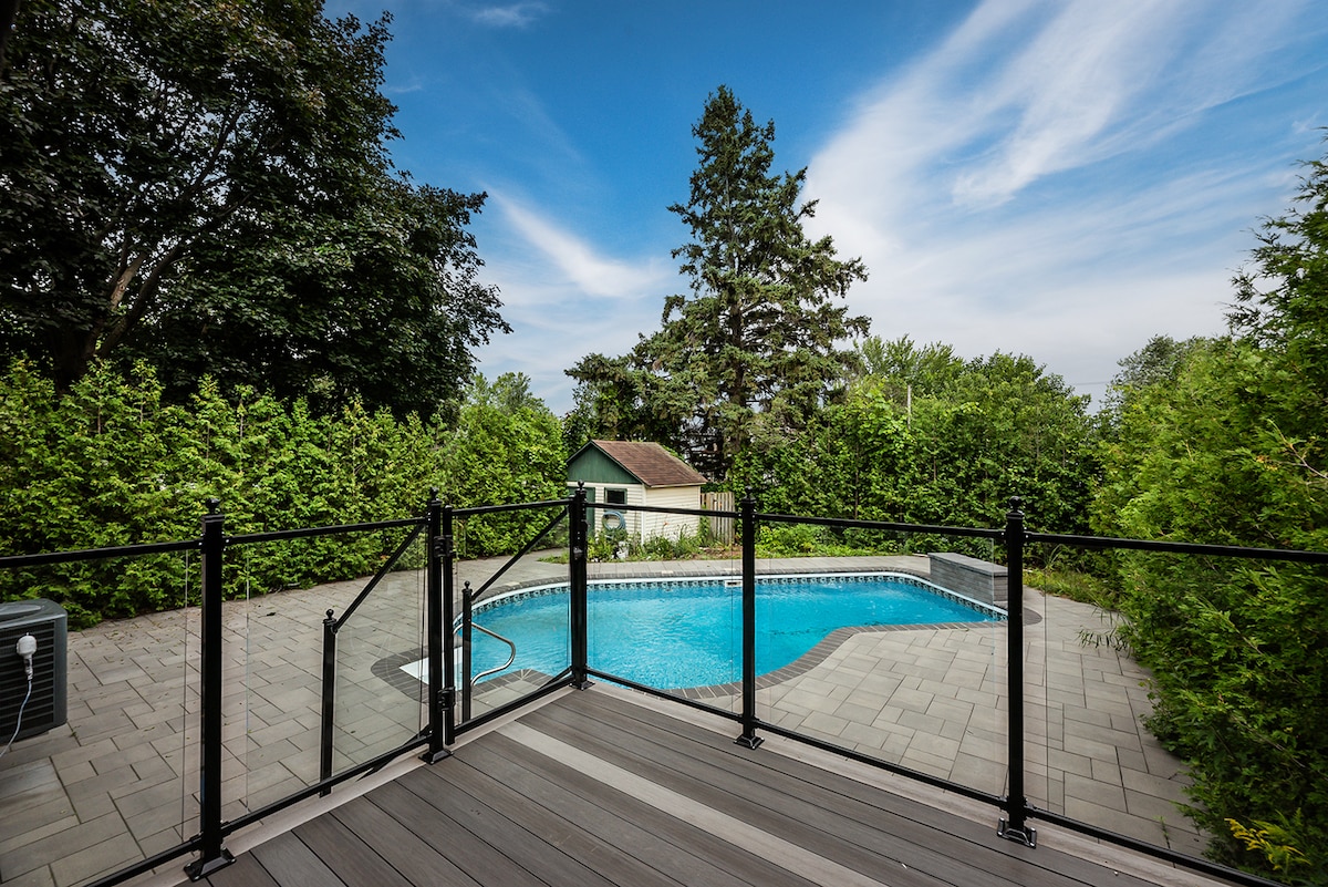 A private pool area is surrounded by lush greenery, creating a secluded atmosphere. The pool features a unique shape and is bordered by smooth paving stones. A safety fence adds an element of security, while a small shed is visible in the background.