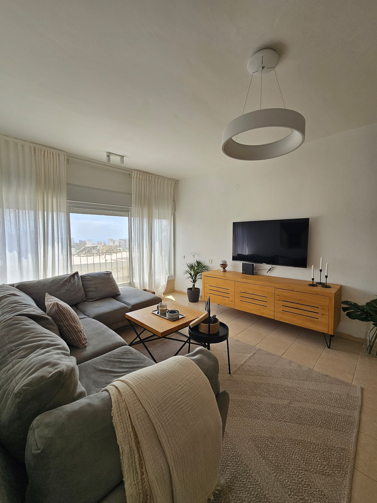 A spacious living area is furnished with a large gray sofa, complemented by a coffee table and decorative accessories. Natural light streams in through a large window, which offers a view of the surroundings. A modern television is mounted on the wall, adding functionality to the space.