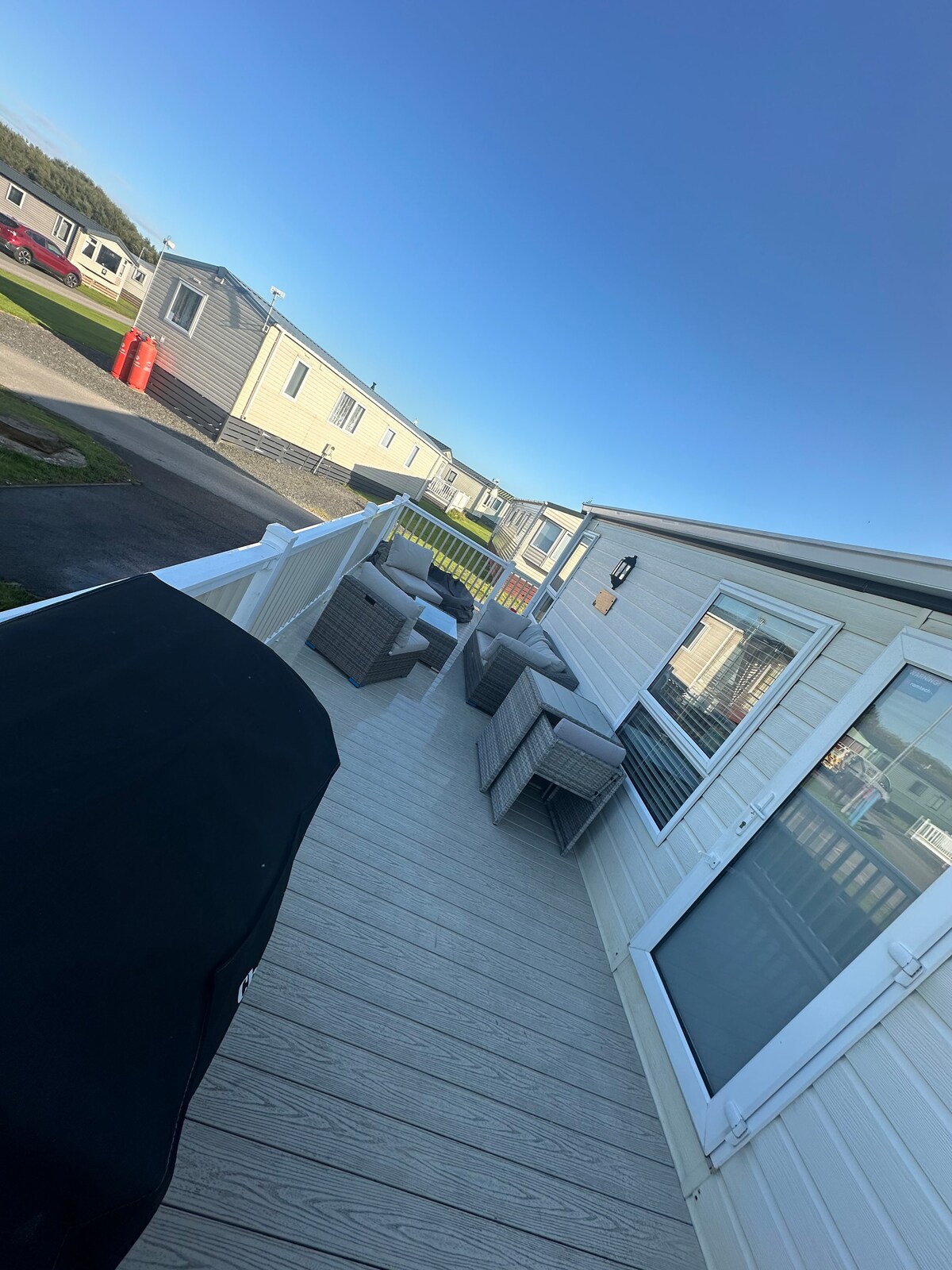 A well-furnished outdoor deck features seating areas with several contemporary chairs and a table, providing a comfortable space for relaxation. A view of neighboring caravans is visible, and a clear blue sky creates a serene backdrop.