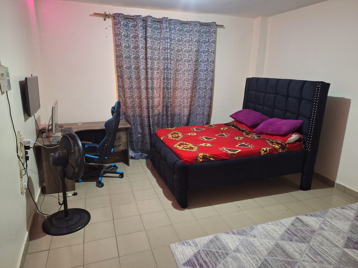 A cozy bedroom features a spacious bed dressed in vibrant red bedding with decorative pillows. A desk with a computer chair sits adjacent to the bed, beside a standing fan. Light emanates softly through patterned curtains, creating a welcoming space.