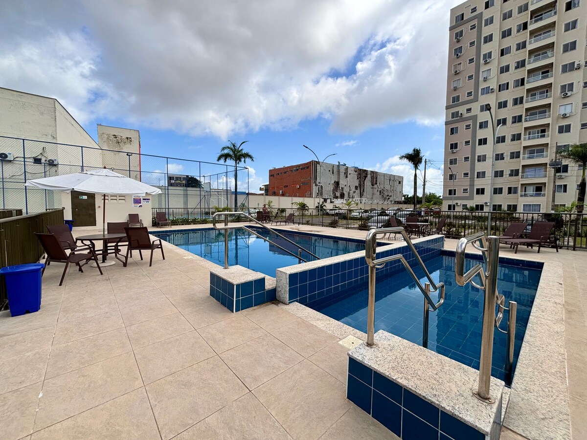 A spacious pool area features a large swimming pool with surrounding lounge chairs and umbrellas. The pool is framed by modern buildings and palm trees, creating an inviting space for relaxation. The tile deck is well-maintained, providing accessibility for all guests.