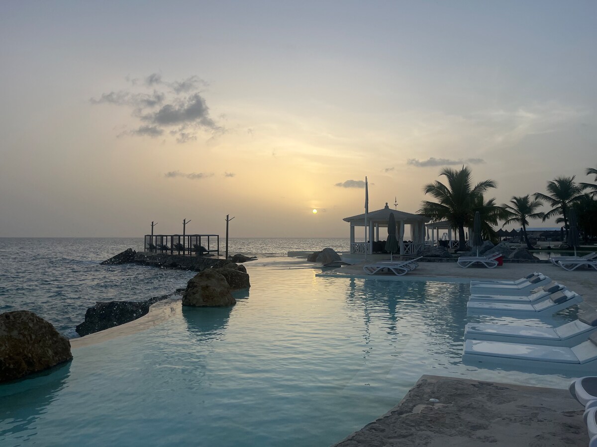 A serene coastal scene features a calm pool reflecting golden sunset hues. Sun loungers are positioned along the water's edge, while palm trees gently sway in the soft breeze. A distant pier extends into the ocean, providing an inviting focal point near the tranquil shoreline.