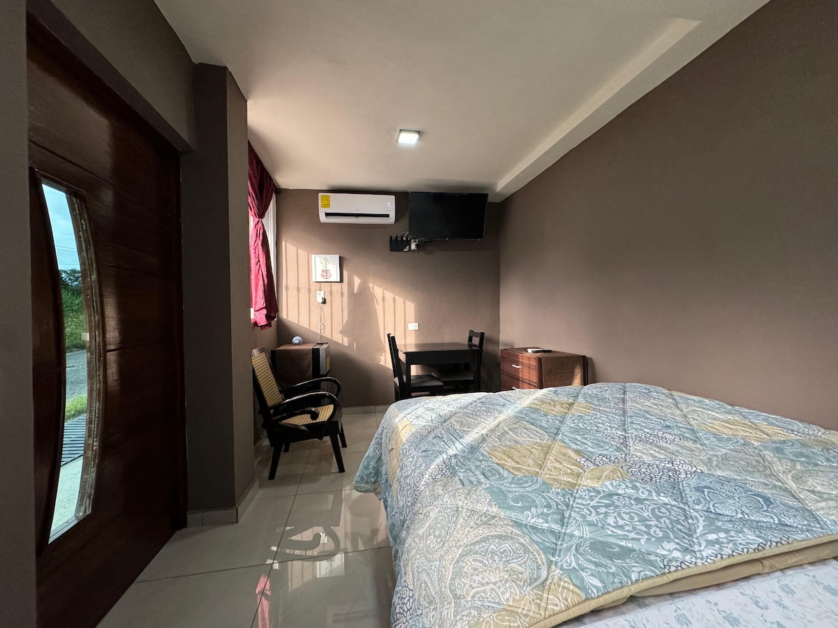 A small bedroom features a spacious bed adorned with a patterned blanket. A television is mounted on the wall, while a wooden desk and chair are positioned near a window. Natural light enters through the window, which is partially covered by a curtain.