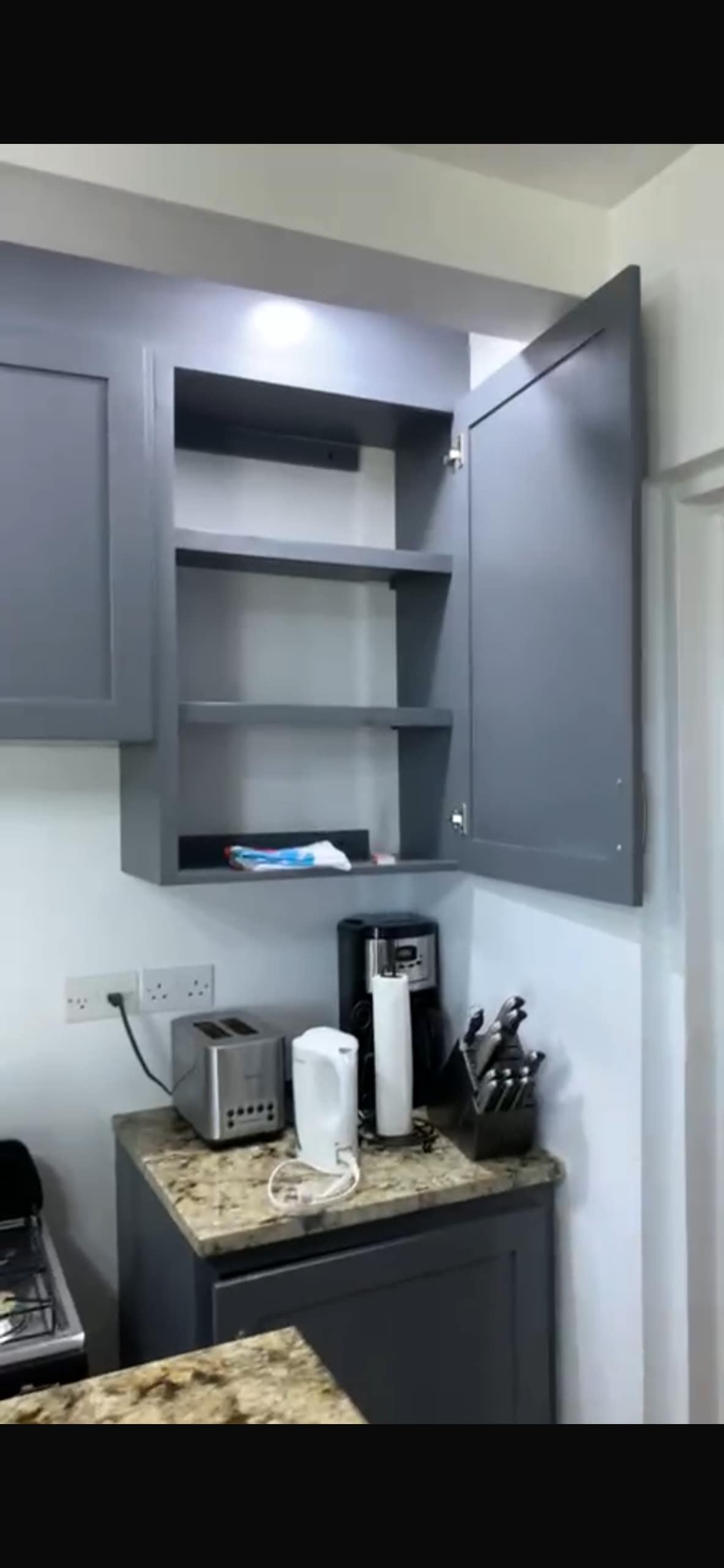 A modern kitchen cabinet with a medium grey finish features open shelving suitable for storing various items. Below, a countertop hosts a toaster and an electric kettle, alongside kitchen utensils organized in a holder.