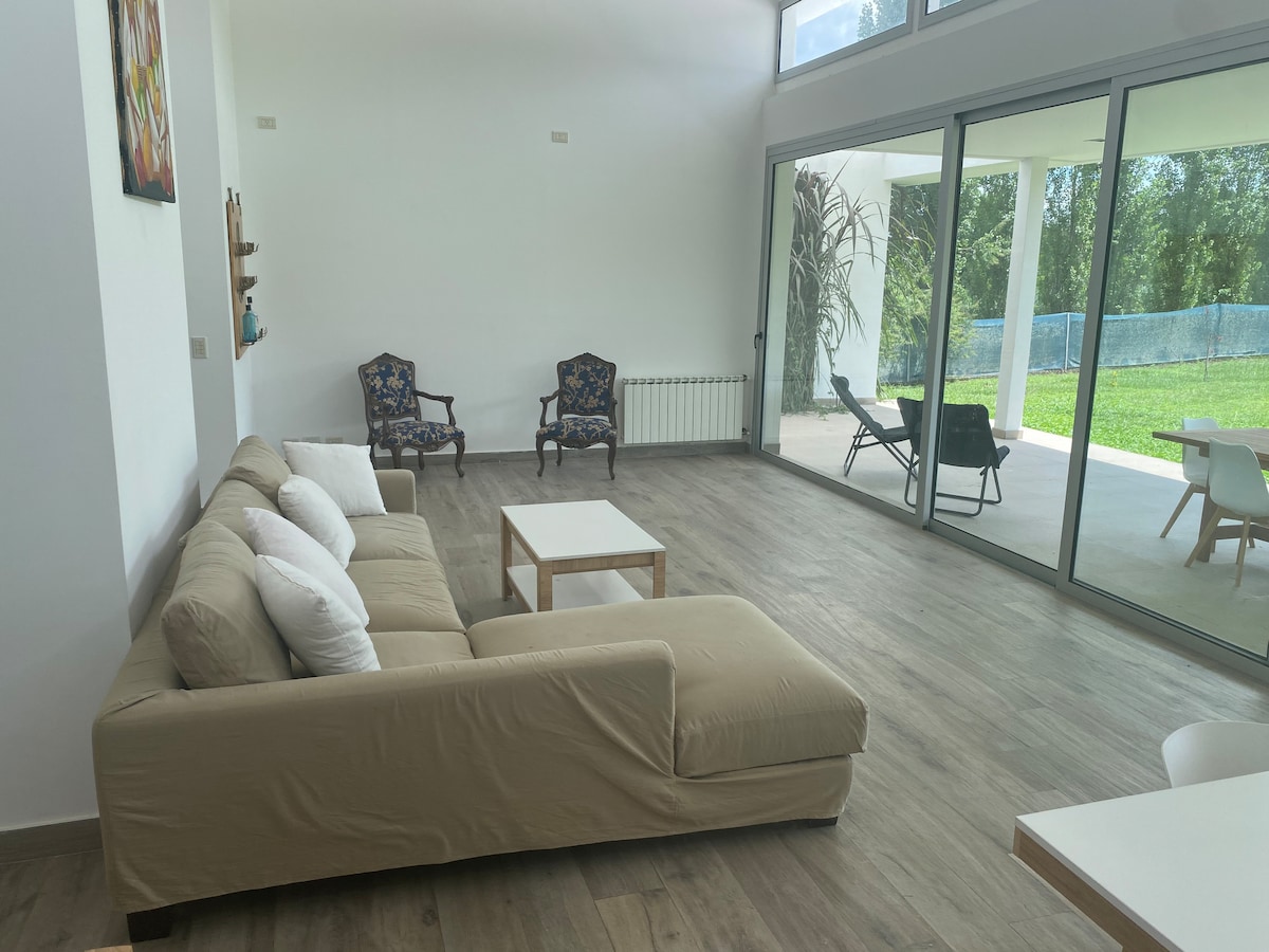 A spacious living area is featured, showcasing a light-colored sectional sofa adorned with white throw pillows. A low coffee table sits in front, while two armchairs are positioned nearby. Large glass doors provide access to a green outdoor space, enhancing natural light in the room.