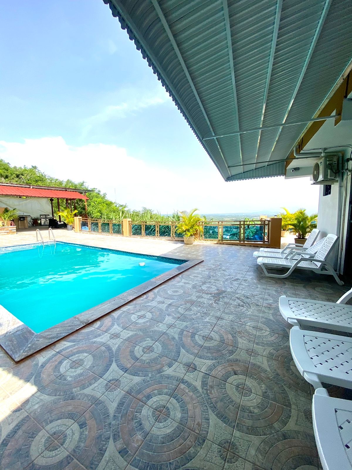 A serene outdoor space is shown with a sparkling pool surrounded by a tiled deck. Lounge chairs are positioned near the water, and lush greenery is visible in the background. The sky is bright and open, contributing to a tranquil atmosphere.