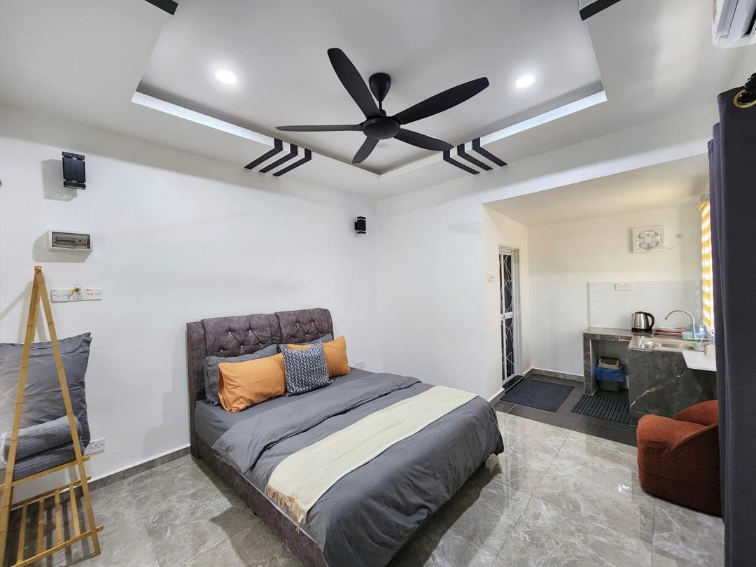 A spacious bedroom features a large bed with soft bedding in neutral tones. A modern ceiling fan is positioned above, complementing the sleek ceiling design. In the corner, a cozy seating area is provided, while a kitchenette is visible in the adjacent space.