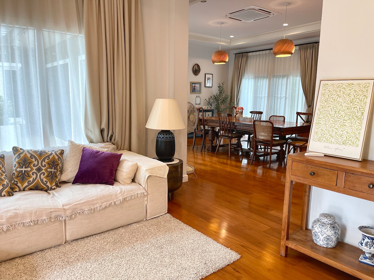 A spacious living area is adorned with a light-colored sofa featuring decorative cushions. Natural light enters through large windows, highlighting the warm wooden flooring. A dining area with a wooden table and chairs can be seen in the background, complemented by soft lighting from pendant fixtures.