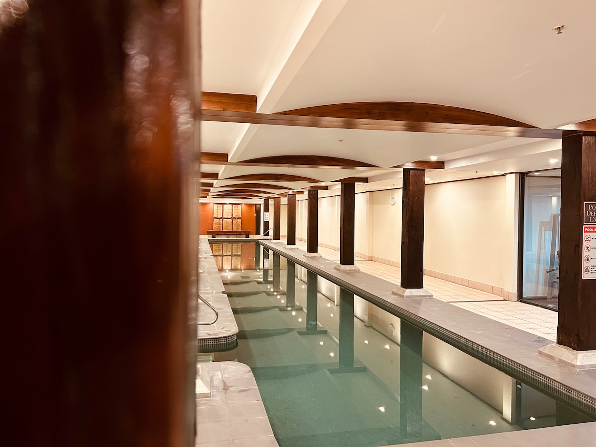 An indoor swimming pool is surrounded by wooden beams and columns, enhancing the open space. Soft lighting reflects on the water's surface, while tiled areas provide access to seating and amenities nearby. The calm atmosphere invites relaxation and enjoyment.