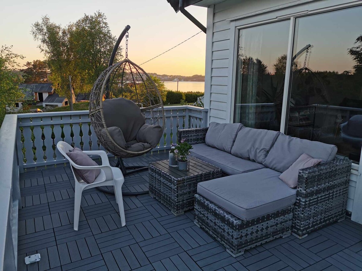 An outdoor terrace is furnished with a spacious sectional sofa and a cozy hanging chair. A small table sits in the middle, complemented by a potted plant. The setting offers views of nature, illuminated by the soft light of dusk.