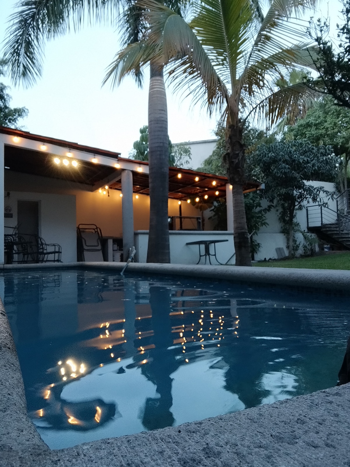 A serene swimming pool is framed by lush greenery and tall palm trees, reflecting warm ambient lighting from overhead string lights. An outdoor seating area is visible in the background, enhancing the inviting and relaxing environment.