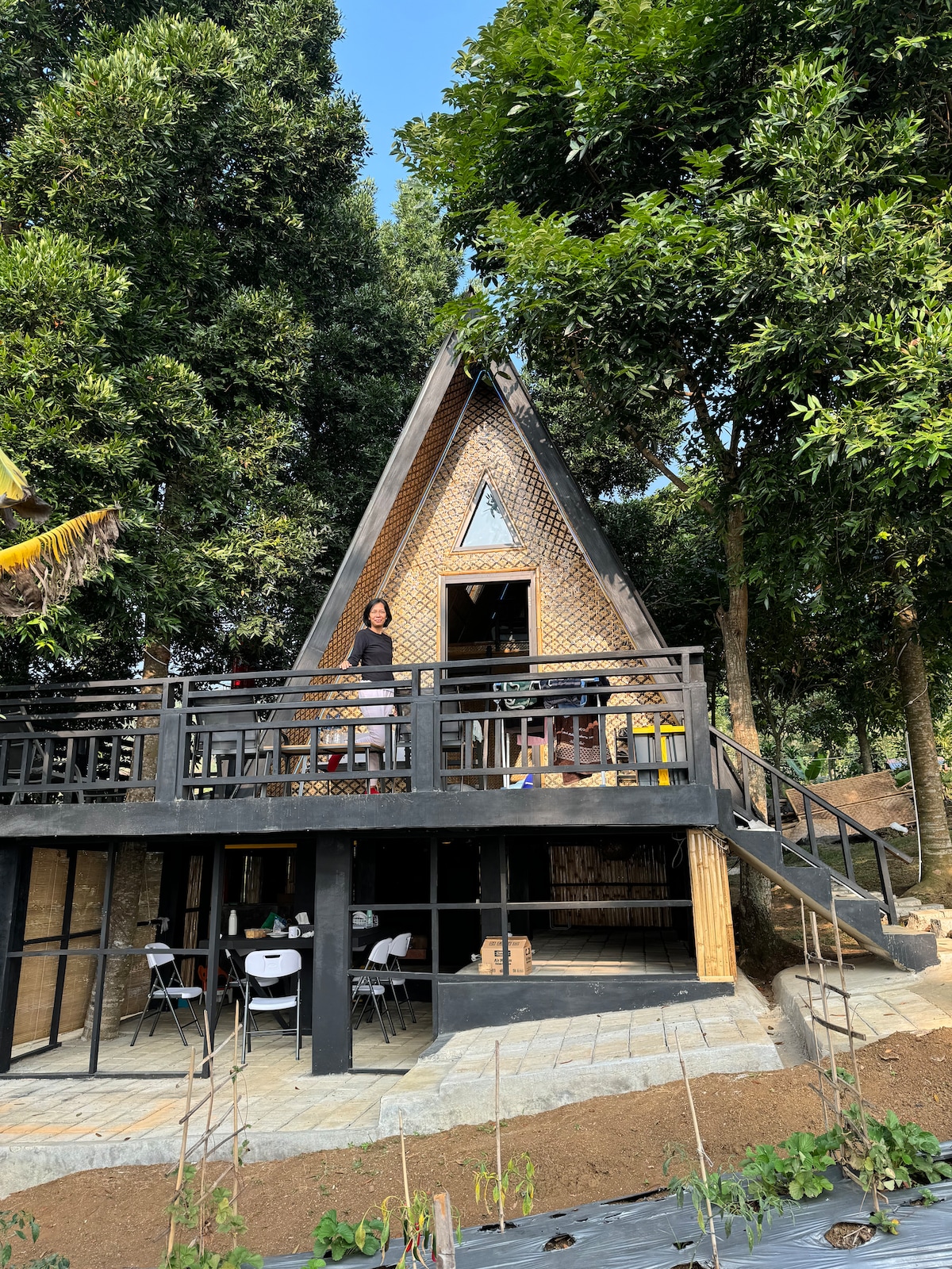 A two-story triangular house features a wooden deck and is surrounded by lush greenery. The exterior showcases a mix of natural materials, while the entrance is accessible via a sloped pathway. Tables and chairs are visible on the deck, providing a space for outdoor relaxation.